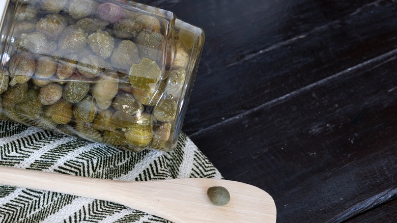 Caper on wooden spoon next to jar of pickled capers