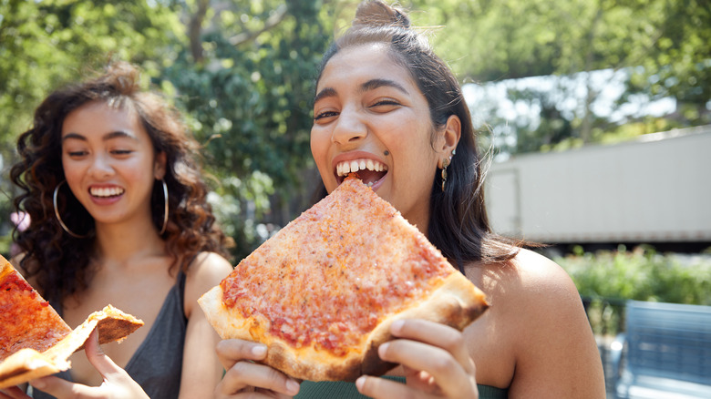 Women smiling and eating NY-style pizza in park