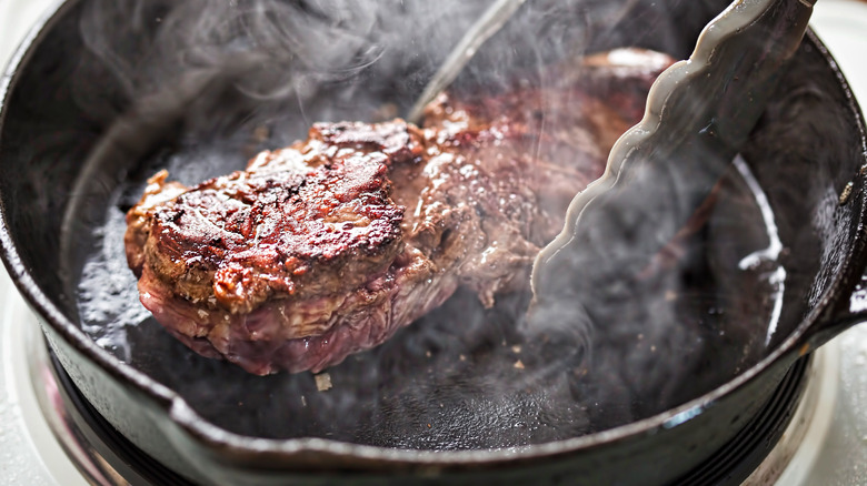 Tongs moving steak in cast iron pan