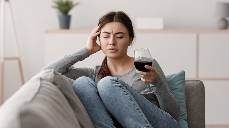 a woman sitting on the sofa with a glass of red wine holding her head in pain