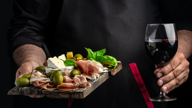 Hands holding a board of sliced prosciutto and glass of red wine