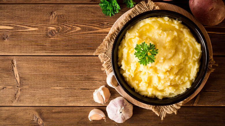 A bowl of mashed potatoes on a rustic wooden table, beside it a bulb of garlic and some opened garlic cloves