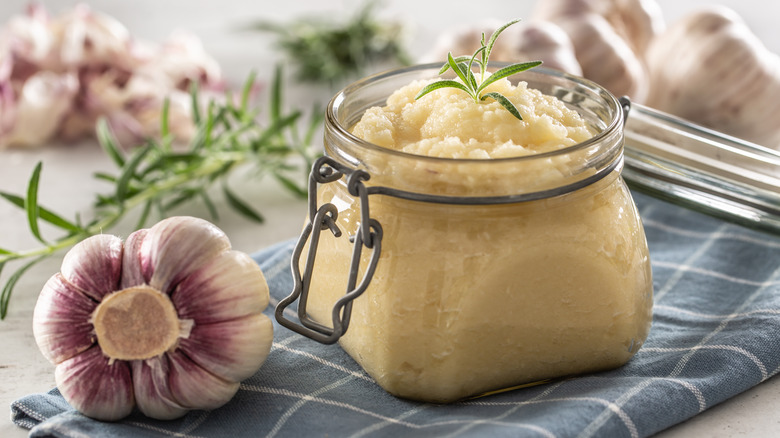 Garlic purée in a small glass jar, lying on a gray and white dish cloth, beside it a full head of garlic