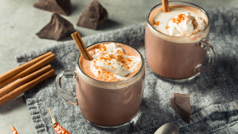 Two mugs of spicy Mexican hot chocolate sit next two cinnamon sticks and dried peppers.