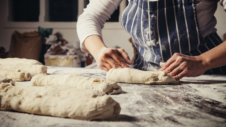 A chef wearing a blue and white striped apron kneads loaves of bread dough into an oblong shape