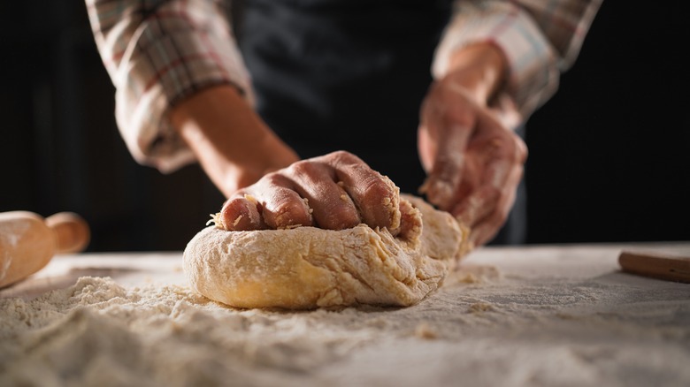 A baker's hands kneads uncooked bread dough on a wooden slab covered in flour