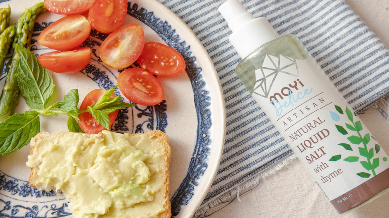 A bottle of liquid salt rests beside a plate of fresh vegetables, herbs, and buttered bread