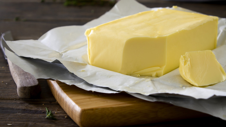 A block of butter sits on a wooden board, with a slice of butter laying adjacent on its side