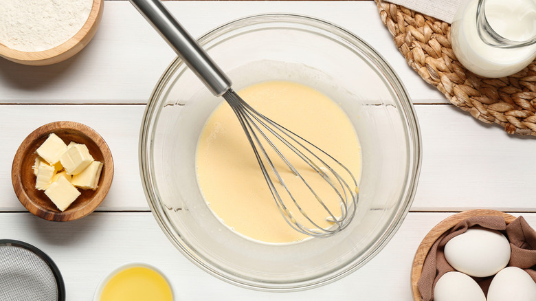 Bowl full of wet crepe batter with whisk and ingredients on white wooden table