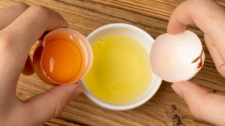 hands holding egg shells, with one holding the egg yolk, with the whites in a ramekin below