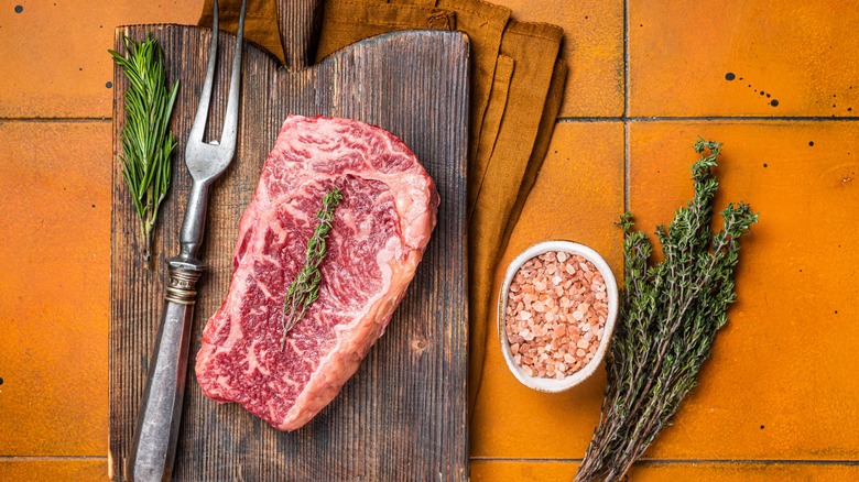 Raw Wagyu striploin steak on board with bowl of salt and herbs