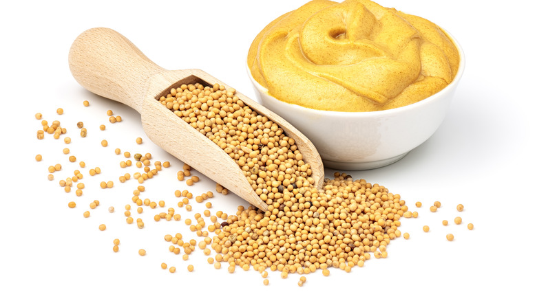Wooden scoop of mustard seeds with bowl of mustard