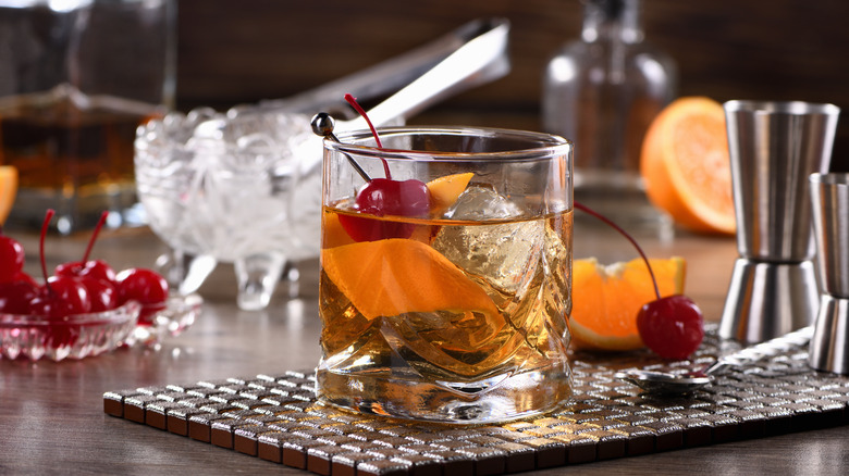Old fashioned cocktail in a glass with cherries, whiskey, and cocktail making kit in the background