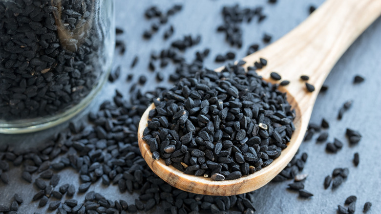 Nigella seeds on a wooden spoon with seeds scattered on a grey table