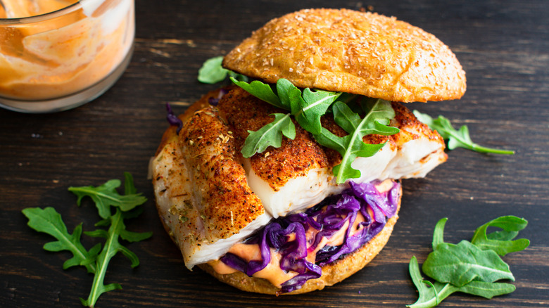 Fried fish sandwich with red cabbage and arugula on a bun with pot of sauce