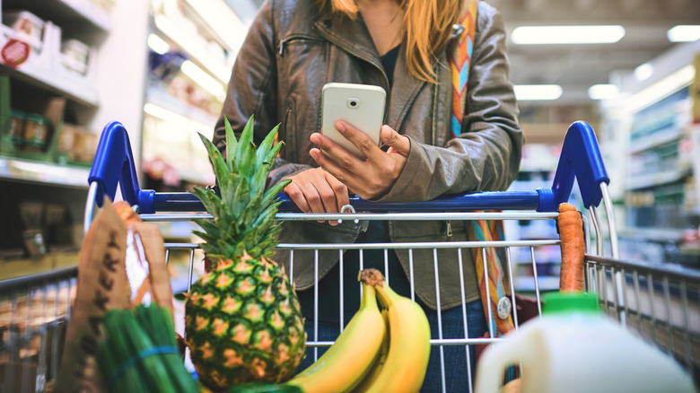 shopper with full grocery cart checking deals on phone