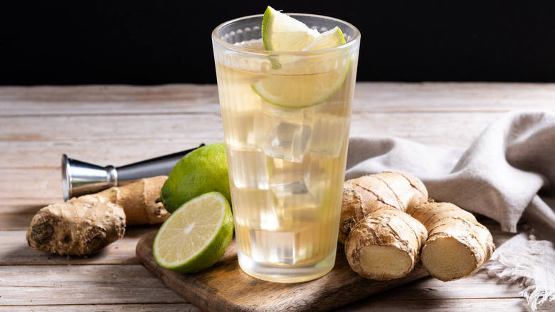 A glass of ginger ale with ice cubes and lime slices sits on a serving board with raw ginger and limes