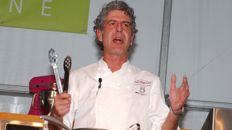 Anthony Bourdain holding tongs in front of a pot while addressing a crowd