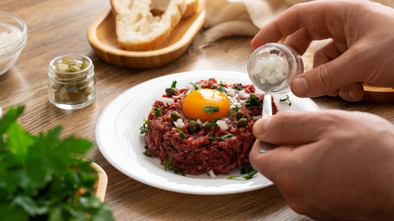 Steak tartare topped with a raw egg yolk on a white plate being seasoned with salt 