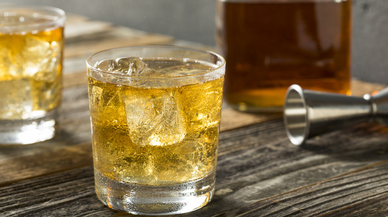 Golden colored, fizzy liquid in short tumbler with a jigger to the right on a flat wooden surface