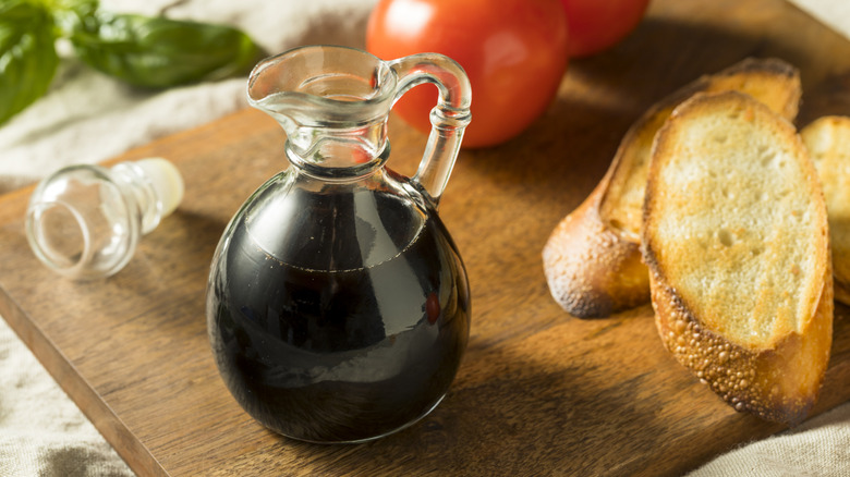 Glass carafe of balsamic vinegar on a wooden cutting board with bread, tomatoes, and basil