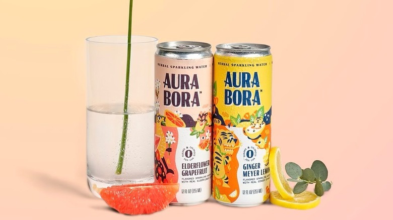 Two cans of Aura Bora seltzer next to a glass with citrus slices and eucalyptus leaves