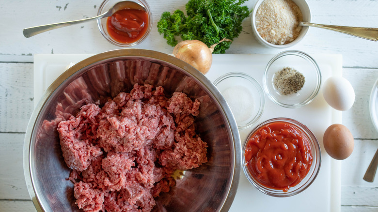 Meatloaf ingredients on a counter, including a bowl of ground beef; vegetables; uncracked eggs; and small dishes of ketchup, breadcrumbs, and seasonings