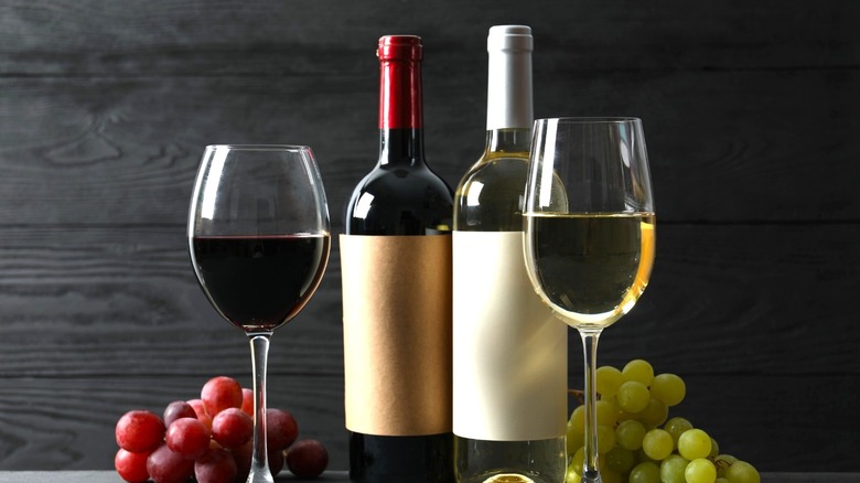 Red and white wine bottles sitting behind wine glasses next to bunches of grapes