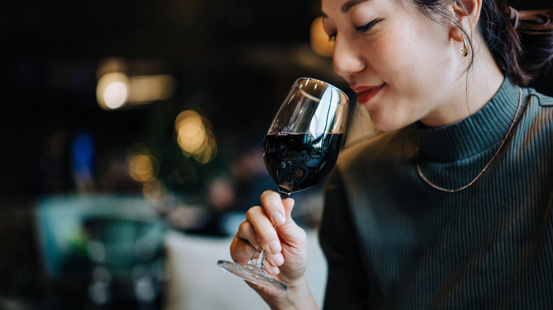 A woman in a green sweater and red lipsticks smells the rim of a wine glass full of red wine