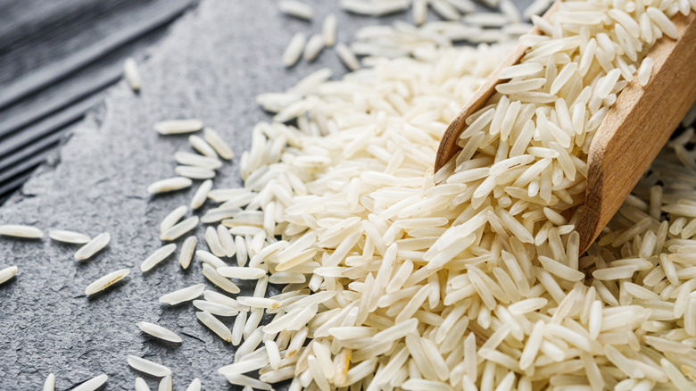 An overflowing wooden scoop of uncooked basmati rice sits on a gray countertop