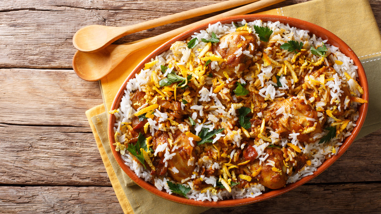 Chicken biryani in an orange bowl is arranged on a yellow napkin with two wooden spools on a wooden table