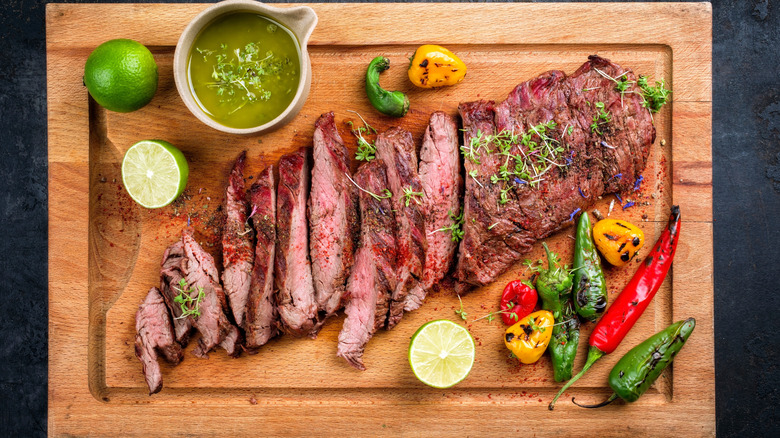 Bavette steak is sliced on a wooden cutting board topped with fresh greens and surrounded by charred peppers, green salsa, and sliced limes