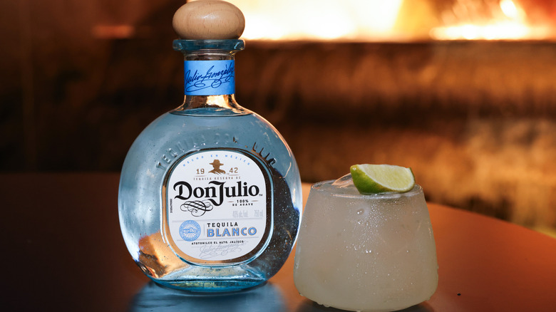 A bottle of Don Julio blanco tequila sits next to a sweating margarita with a lime wedge garnish