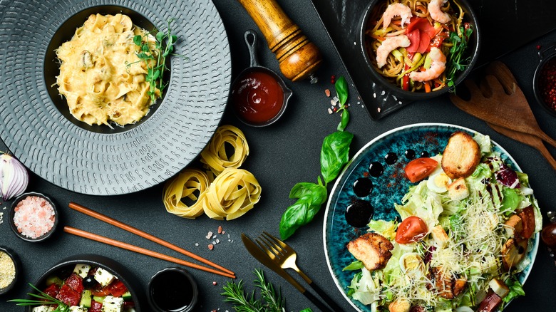 Variety of meals including creamy pasta dish, salad with balsamic, and shrimp pasta dish on a gray table. Additional pasta, sauces, seasonings, and utensils all surrounding the plates.