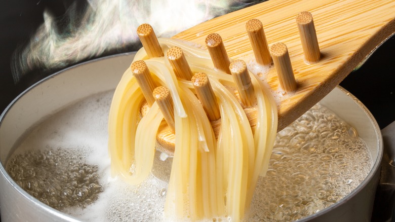 Pasta being taken out of boiling liquid with wooden ladle. Steam is behind the white pot, all on a dark background.