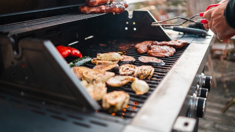 Man with tongs at a grill cooking various meats and vegetables