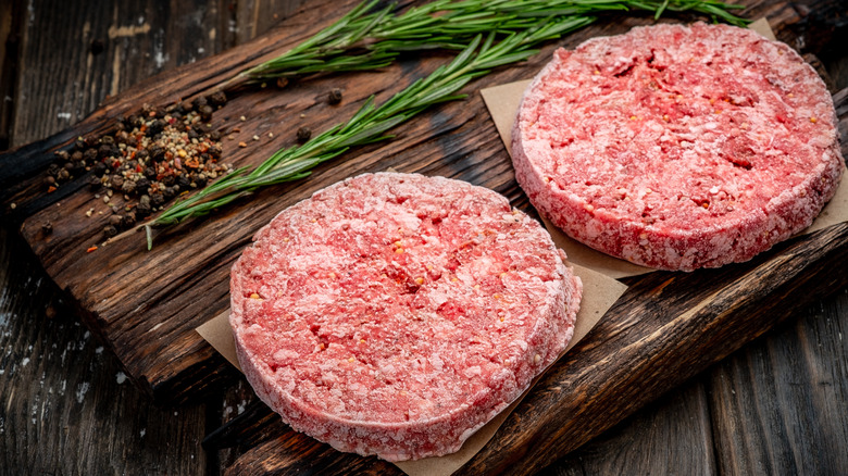 Frozen raw burger patties on a wooden board with herbs