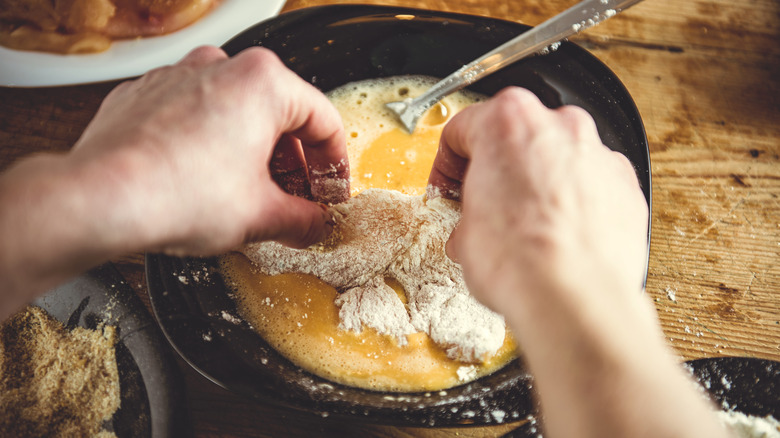 Hands dipping dredged chicken in an egg wash