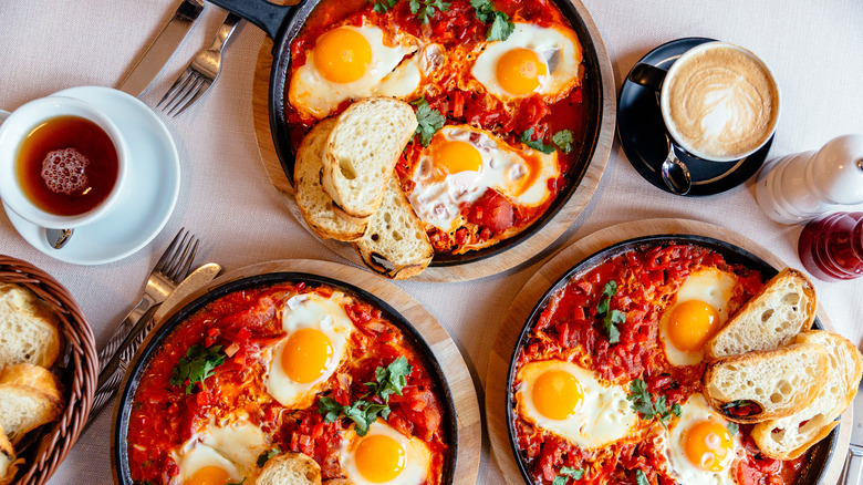 Breakfast spread with three cast iron pans of shakshuka and eggs with toast, cappuccino, and tea