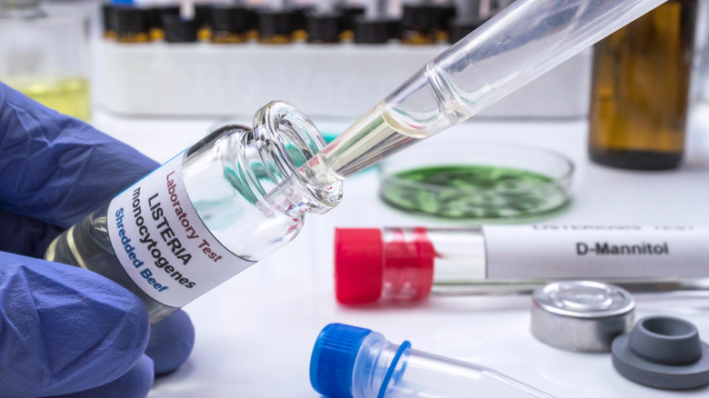 A gloved hand holding a test tube with the label "Listeria monocytogenes"