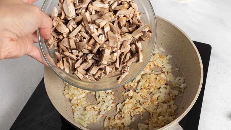 Chopped mushrooms being added to a pan with cooked onion.