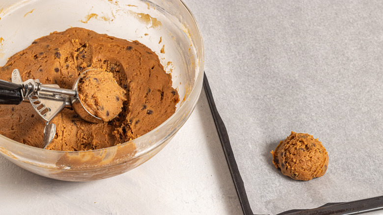 Cookie dough scoop inside a bowl with cookie dough and one cookie dough on a baking tray 
