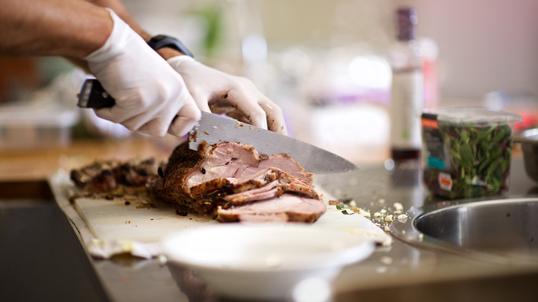 Roast beef being sliced on a white plastic cutting board with glove covered hands