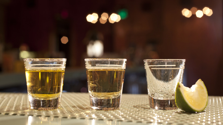 Three tequila shots sit on a bar, two gold and one silver, next to a wedge of lime