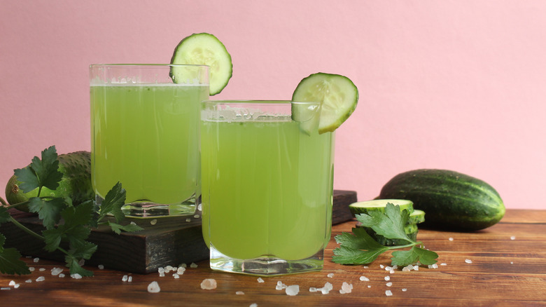 Two cucumber cocktails sit on a wooden table against a light pink background