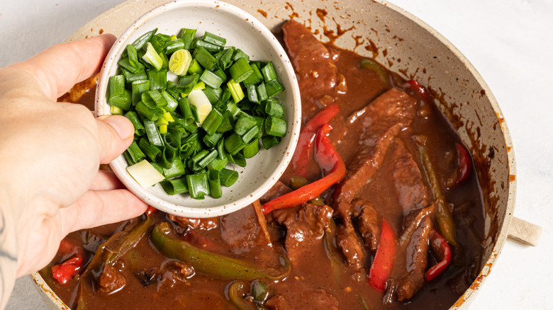 Adding green onion to a skillet with pepper steak in a brown sauce