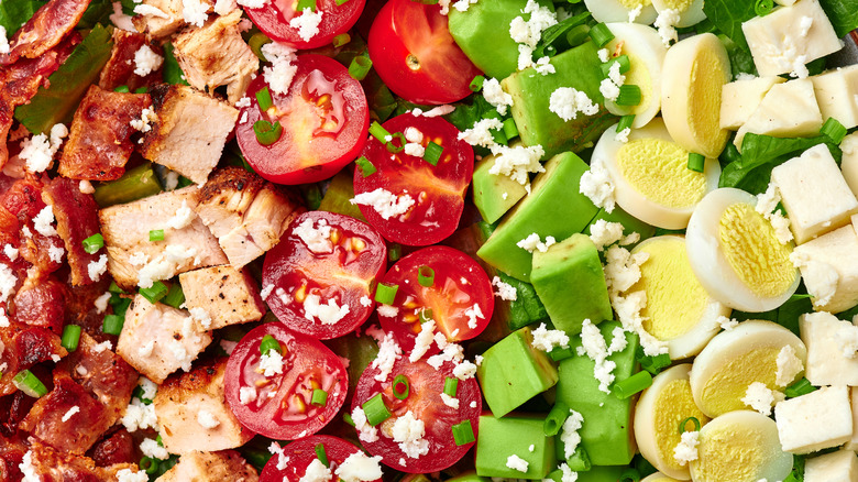 Close up of cooked, chopped bacon, chicken, tomatoes, avocadoes, eggs, and cheese in rows