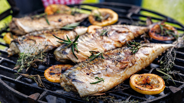 Four headless fish on a grill topped with fresh herbs and lemon slices