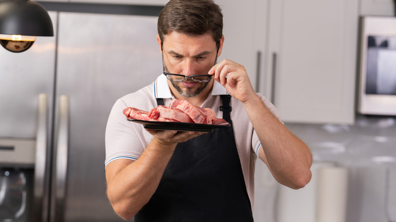 Person in kitchen lifts their glasses to look at raw steak on a plate