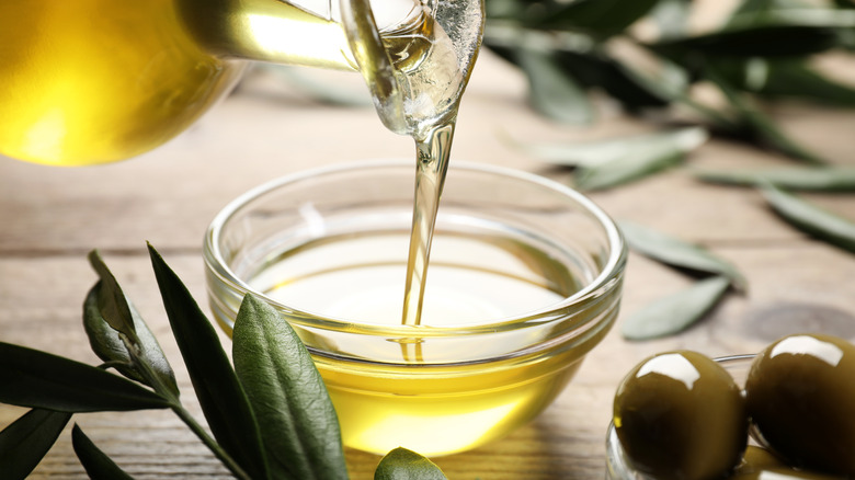 Olive oil pouring from a bottle into a bowl, surrounded by olives and olive leaves.